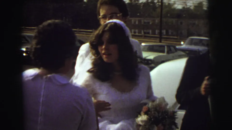 The bride, holding a small bouquet, stands beside the car with relatives around her in the 1972 East Lansing wedding scene.