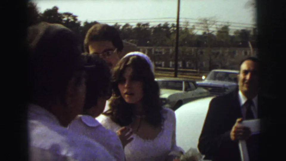 In 1972 East Lansing, a bride in a veil and a groom in a light suit stand with guests near a white limousine and parked cars.