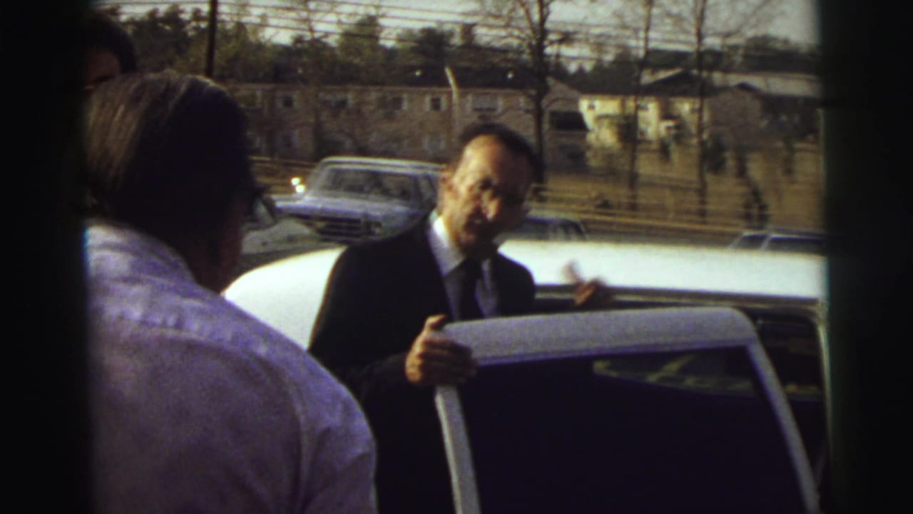 Man in a dark suit opens a white limousine door as guests stand nearby.