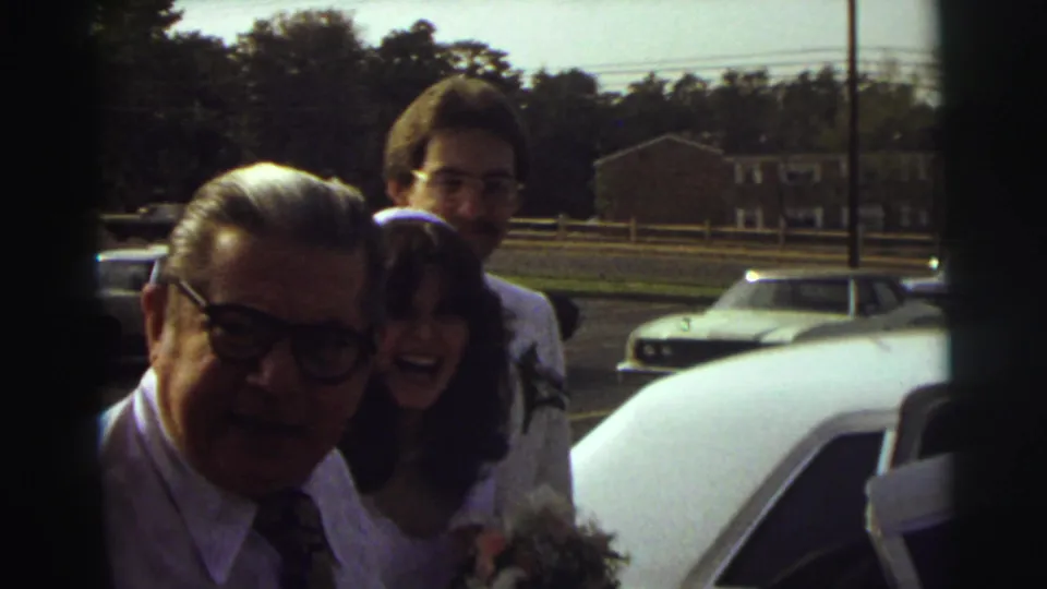 At the East Lansing wedding send-off, an older guest smiles close to camera as the bride and groom stand near the white limousine.