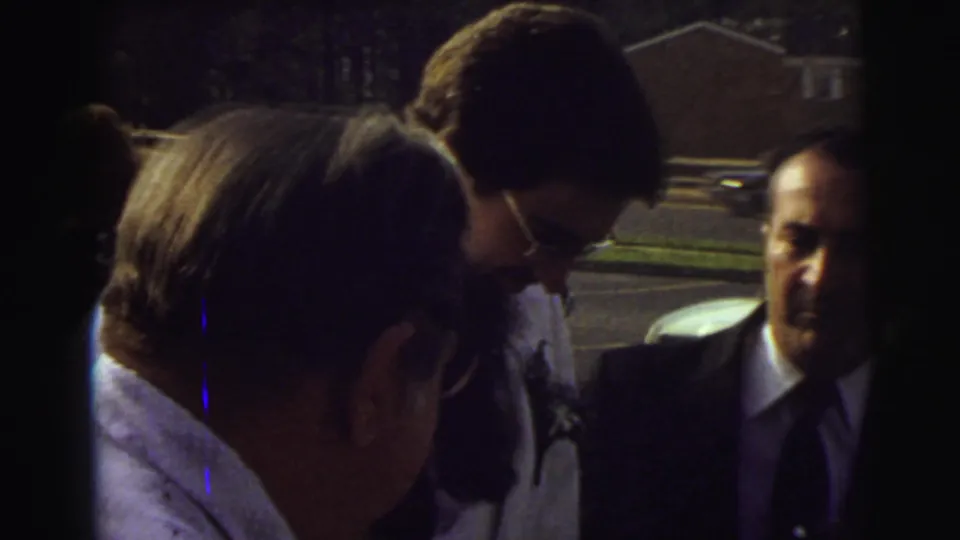 The bride and groom lean toward the open limousine door while suited guests stand nearby in the 1972 East Lansing wedding footage.
