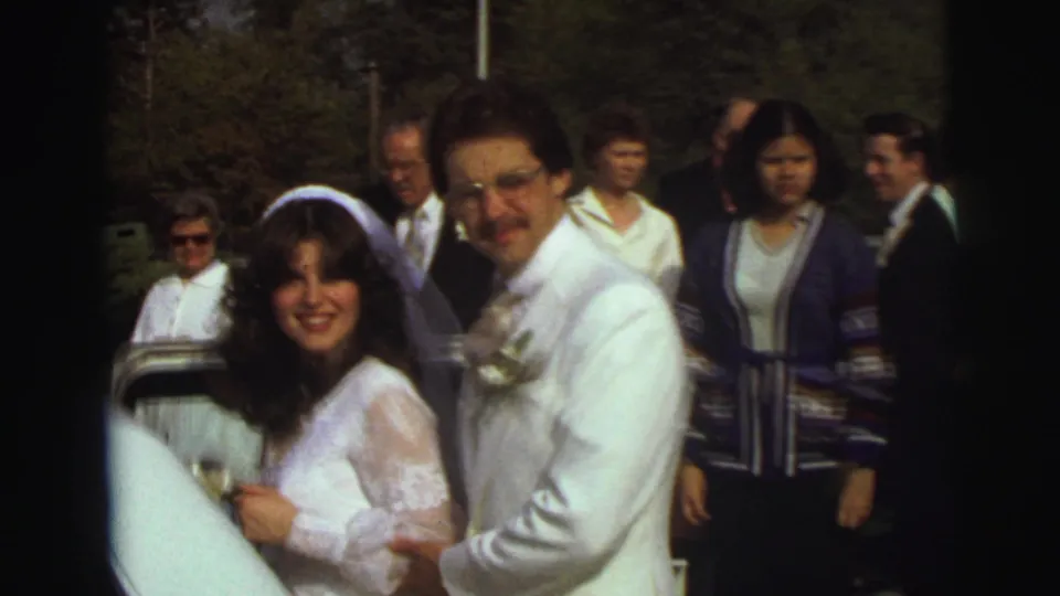 The bride and groom smile close to the white limousine during an outdoor 1972 East Lansing wedding moment, with guests visible behind them.