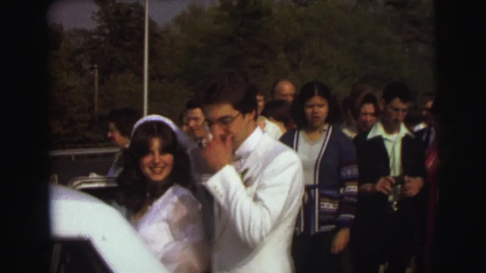 The bride smiles toward the camera while the groom adjusts his face or glasses beside the limousine, with wedding guests gathered behind them.