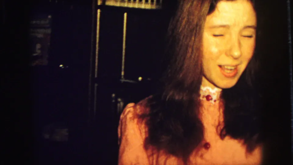 Young woman in a pink dress with a white collar stands in a dim 1973 Superior, Wisconsin home interior.