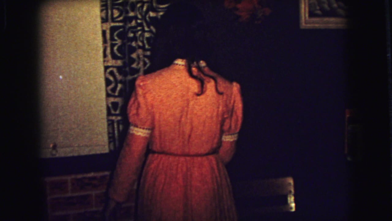 Woman in orange dress seen from behind near a patterned curtain in a dim room.