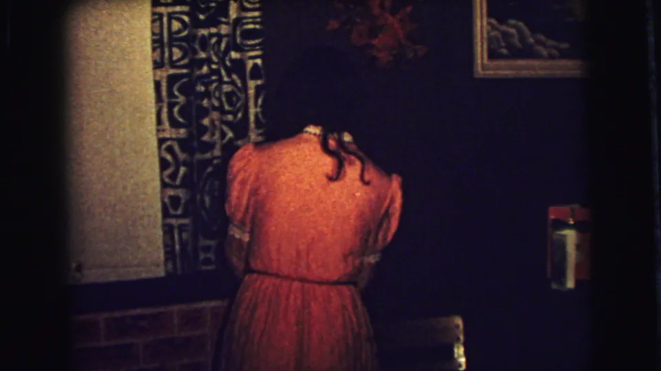 The woman remains turned away in a dim Superior, Wisconsin interior, leaning near patterned decor and a dark wall.