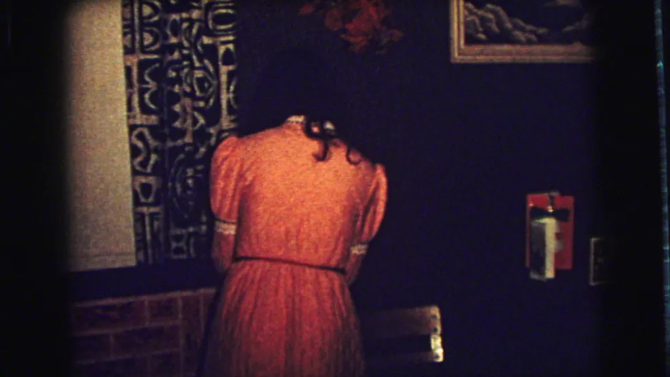 A woman in an orange dress bends forward near a patterned curtain, brick accent, and wall fixture in the 1973 interior.