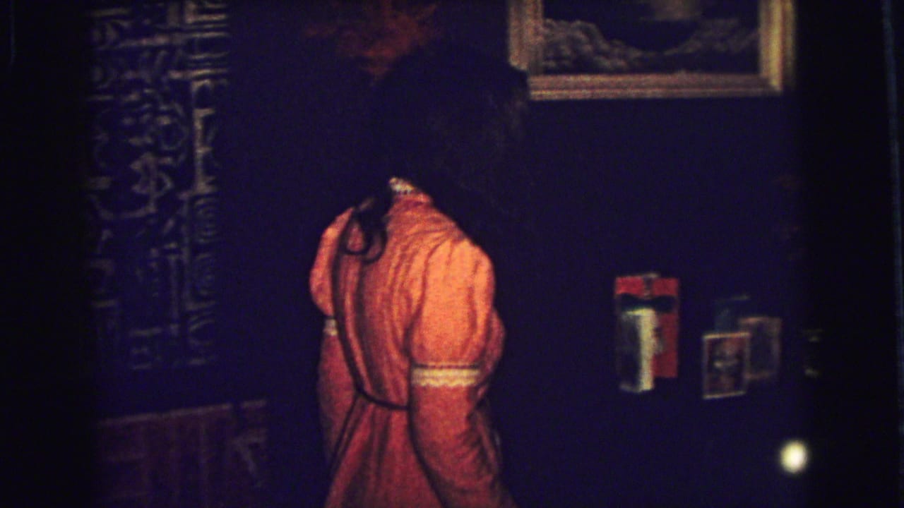 Young woman in an orange dress walks past dark walls and patterned drapes.