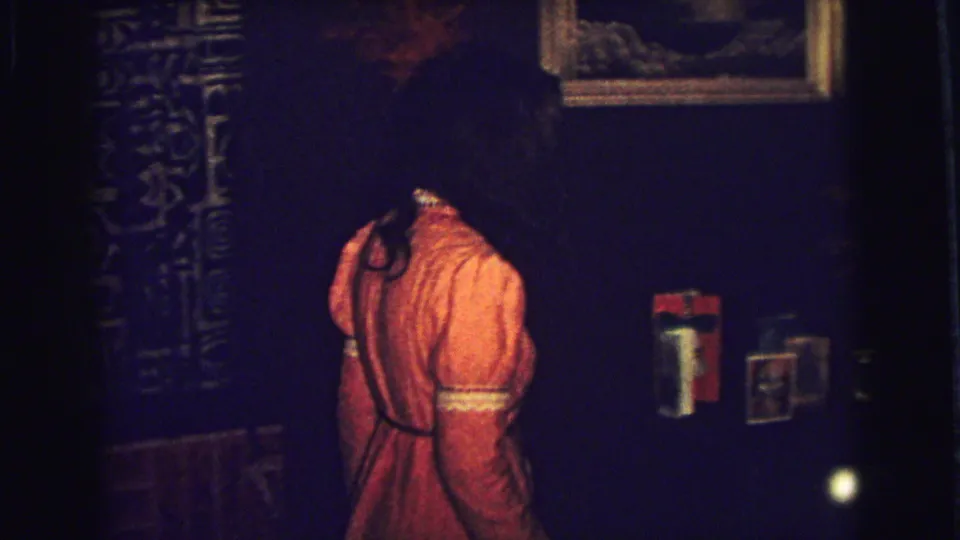 In a 1973 Superior, Wisconsin home-movie frame, a young woman in an orange dress moves through a dim private room.