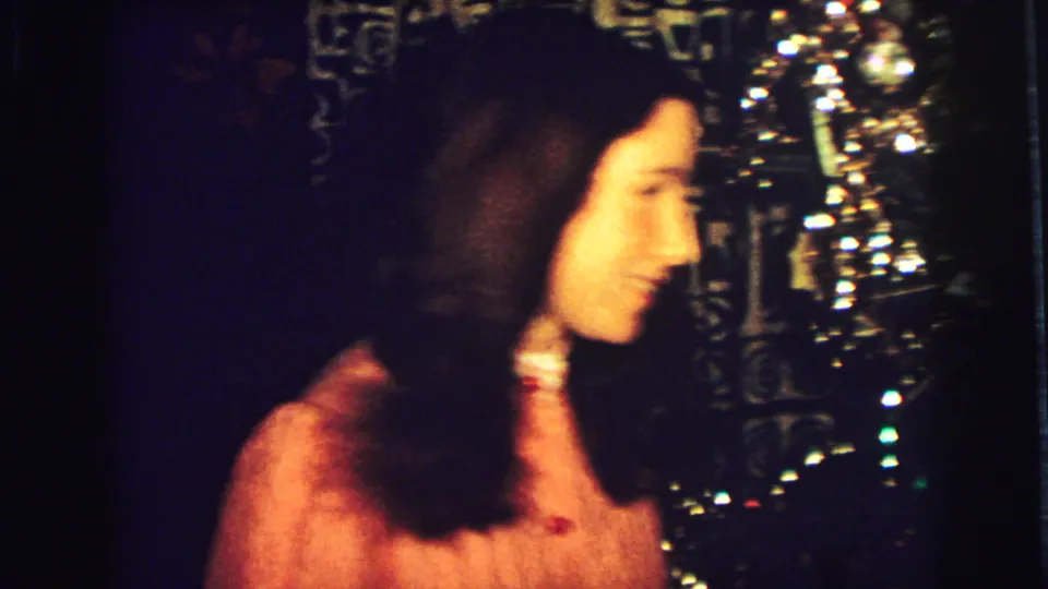 The woman in the orange dress turns in profile and smiles inside the dim 1973 Superior, Wisconsin home interior.