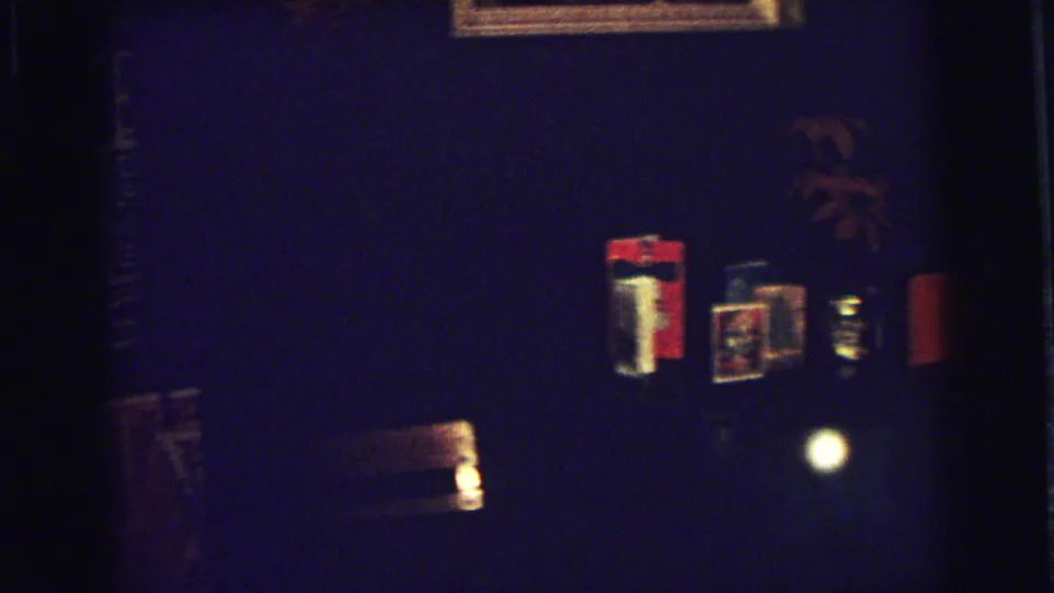 A low-light home-movie frame shows small wall objects and a partially visible person in a dark 1973 Superior, Wisconsin room.