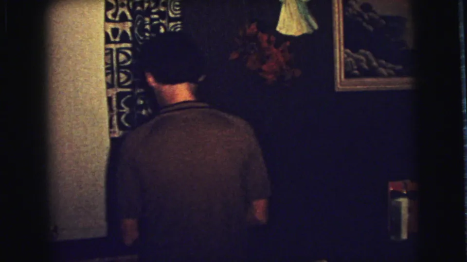A young man in a dark shirt faces away near patterned drapes and framed wall art in the 1973 Superior, Wisconsin interior.