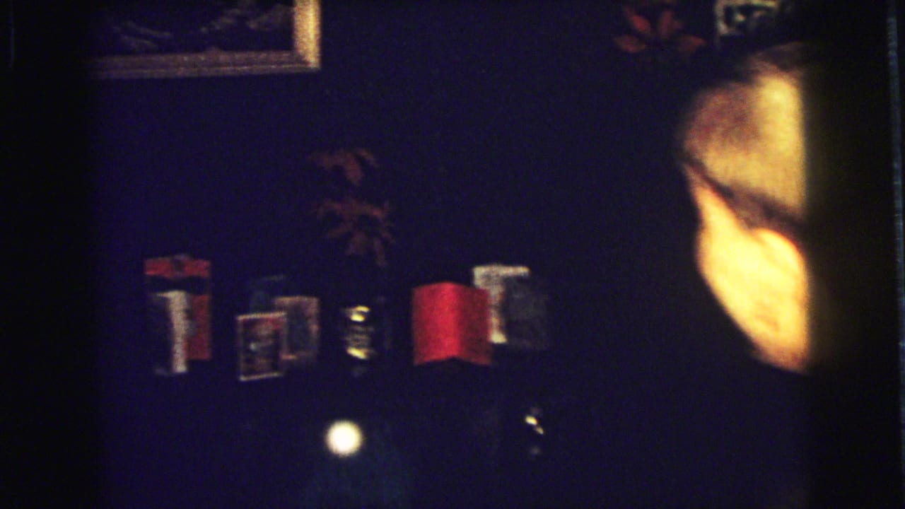 1973 Superior, Wisconsin home-movie frame showing a child in a dark coat near a dim mantle area with a red stocking and wall decor.
