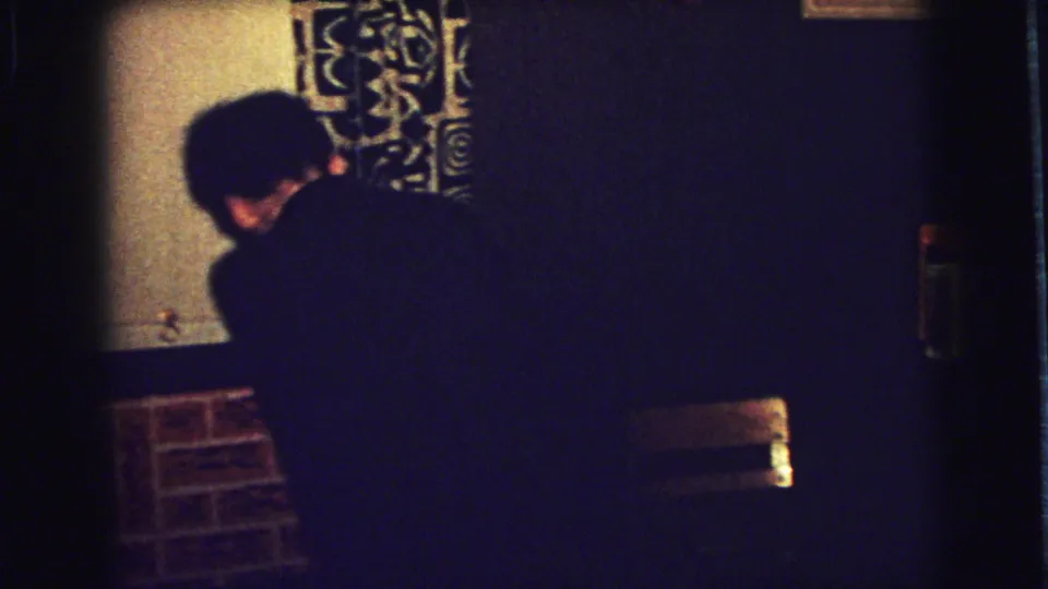 1973 home-movie frame from Superior, Wisconsin showing a child seen from behind reaching near a mantle with patterned curtains behind.