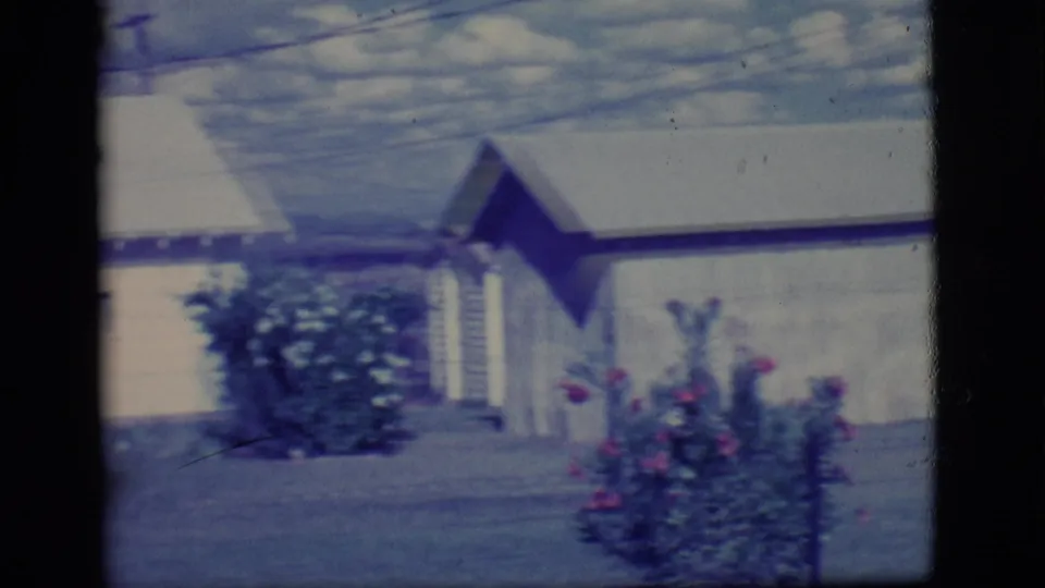 1968 Ely, Nevada home-movie frame showing a light-colored single-story house, shrubs, flowers, and a quiet yard under soft 8mm color.