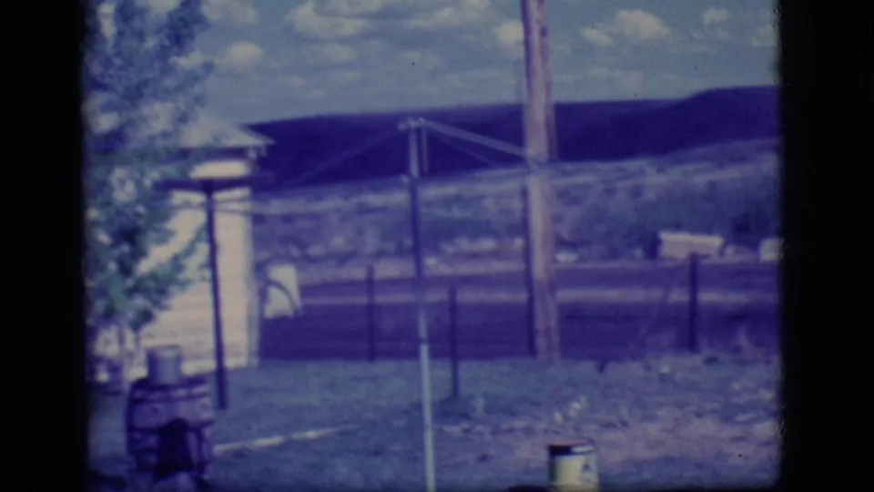 1968 Ely, Nevada frame showing a fenced yard with a utility pole, small yard structures, grass, and low hills in the background.