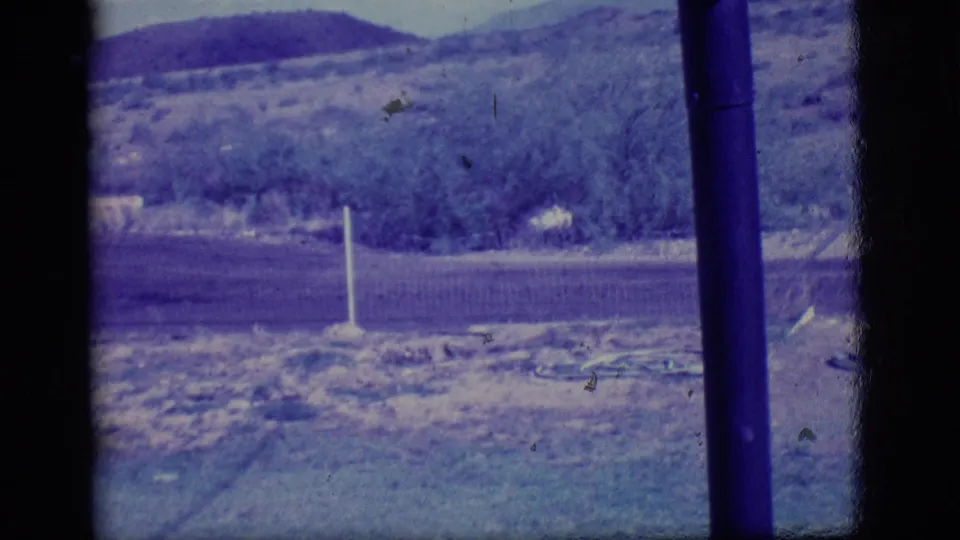 1968 Ely, Nevada home-movie frame showing a dry roadside field, brush, a white post, and hills divided by a dark window bar.