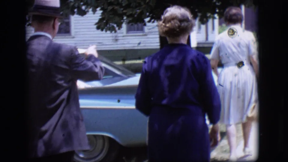 In Des Moines, Iowa in 1966, a suited man and two women in dresses walk toward a blue car outside a house.