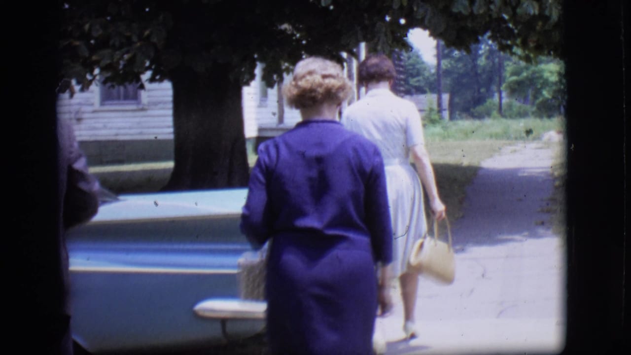 In 1966 Des Moines home-movie footage, two women in dresses approach a light blue tailfin car near a residential sidewalk.