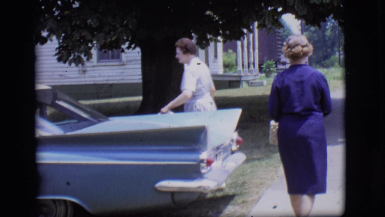 A woman in a white dress opens the door of a light blue tailfin car as another woman in purple stands on the sidewalk in Des Moines.