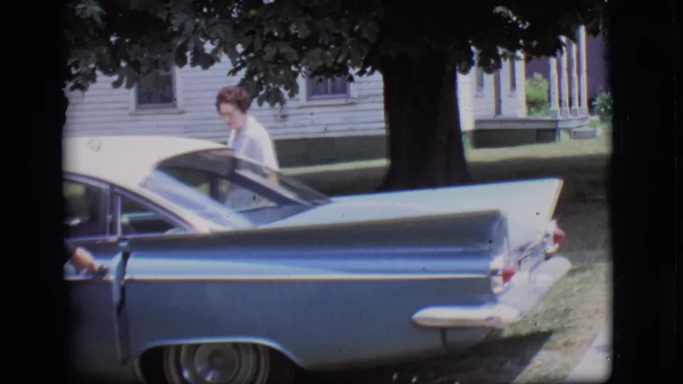 A woman in a white dress stands beside the open side of a light blue tailfin car on a shaded residential street in 1966 Des Moines.