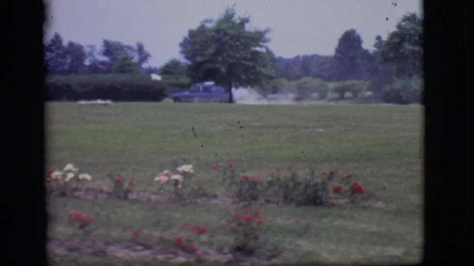 The light blue car is seen farther away on a residential street beyond a lawn and rose garden in Des Moines home-movie footage.