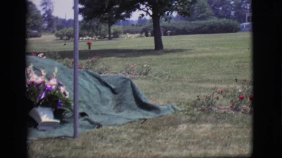 Prepared gravesite in a Des Moines cemetery, with a green covering beside floral arrangements and lawn.