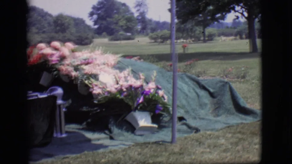 1966 Des Moines cemetery frame showing floral arrangements beside a green-covered gravesite and open lawn.