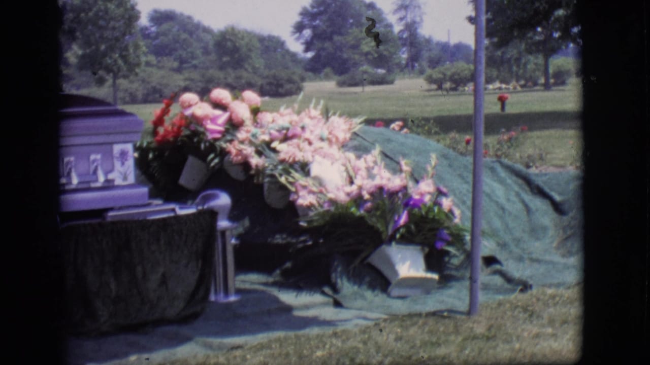1966 Des Moines cemetery view with a purple casket, flower arrangements, green grave covering, lawn, and trees in the background.