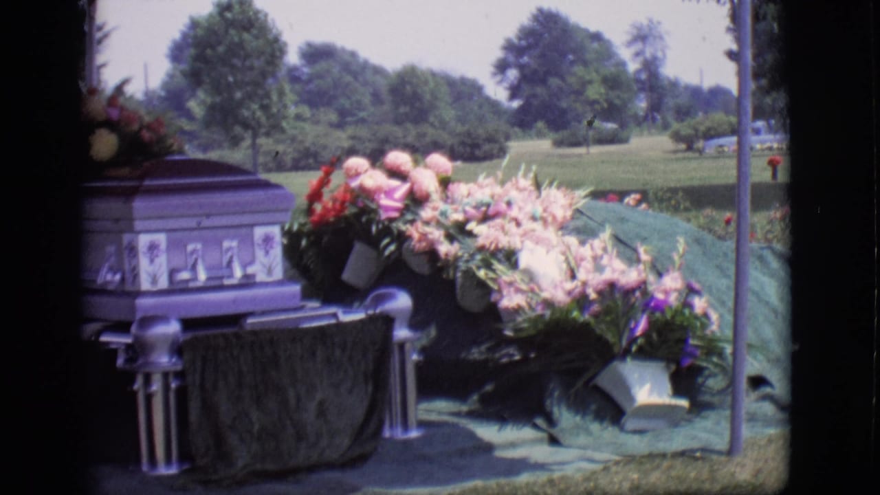 Purple casket beside a covered grave in Des Moines, Iowa, with pink and red flowers, grass, trees, and a distant vehicle visible.