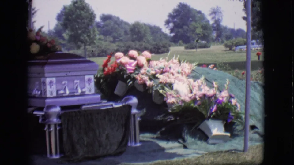 Purple casket beside a covered grave in Des Moines, Iowa, with pink and red flowers, grass, trees, and a distant vehicle visible.