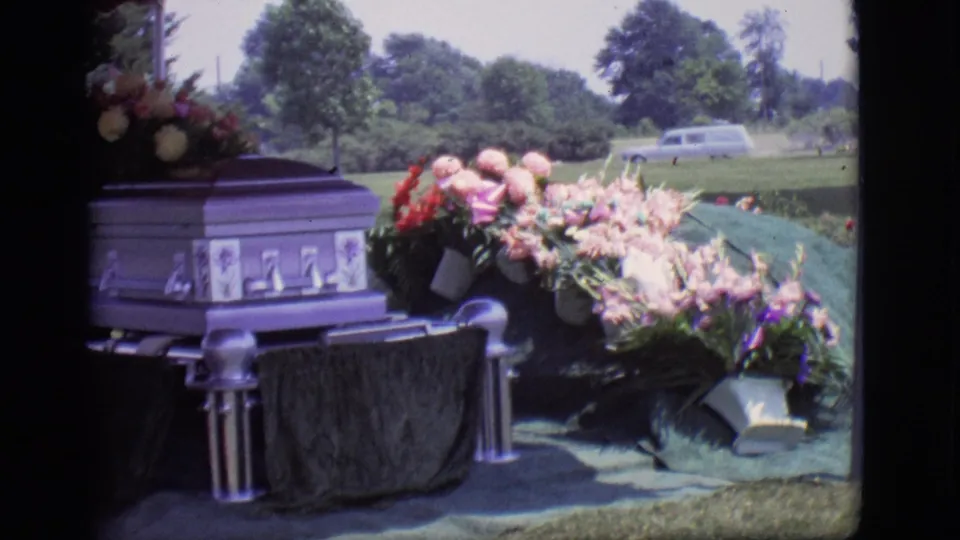 1966 Des Moines funeral frame showing the purple casket, floral sprays, green-covered grave, cemetery lawn, trees, and a distant hearse-like vehicle.
