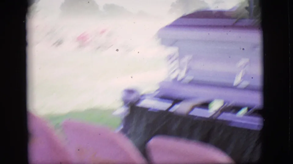 Motion-blurred 8mm frame from the Des Moines funeral scene, with the purple casket and pale floral area still partly visible.