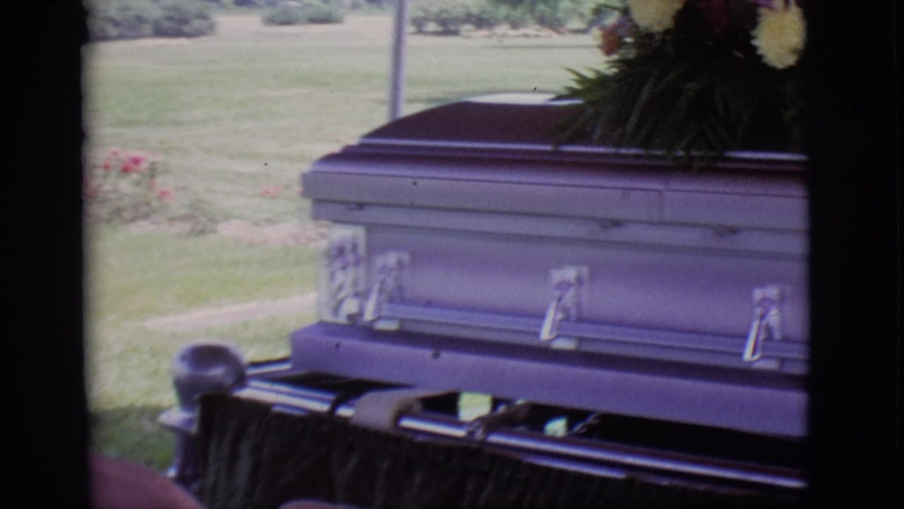 1966 Des Moines home-movie frame showing a casket on a hearse with flowers, grass, and trees behind it.