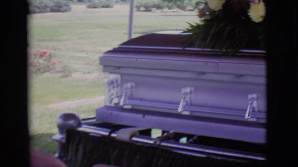 1966 Des Moines home-movie frame showing a casket on a hearse with flowers, grass, and trees behind it.