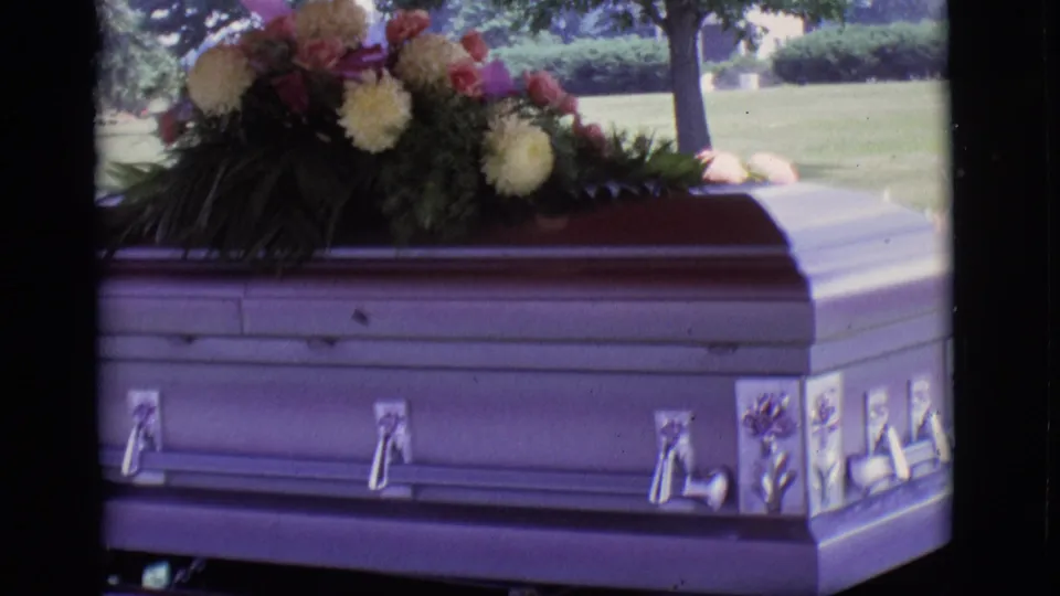 Home-movie frame of the casket corner, floral spray, and hearse support in an outdoor Des Moines funeral scene.