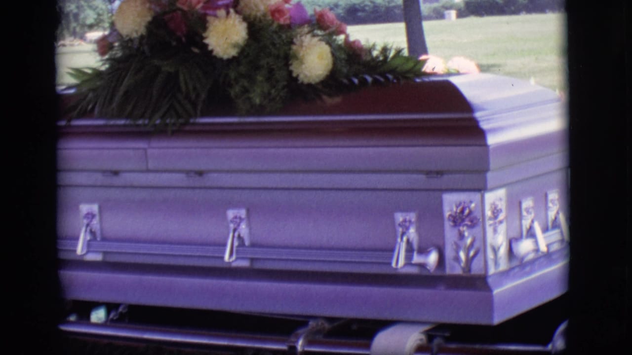 1966 Des Moines home-movie frame showing a lavender casket with ornate handles and yellow and pink flowers resting above it.