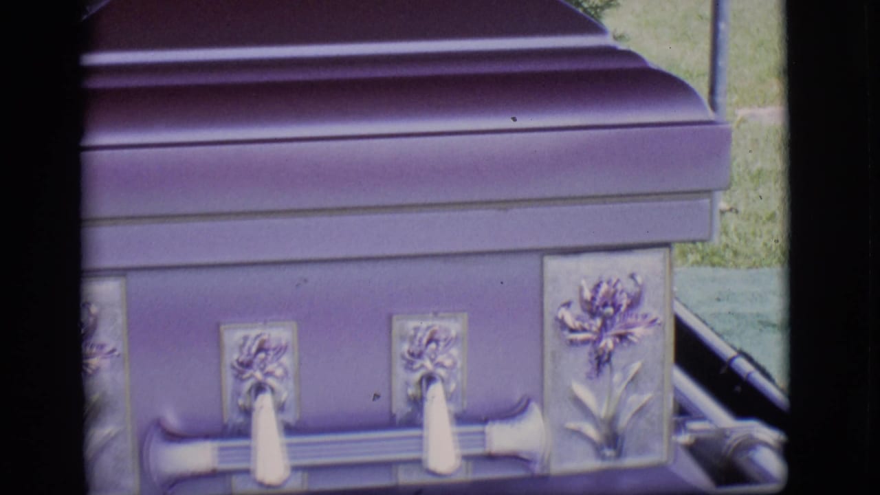 Close 1966 Des Moines frame of a lavender casket side, showing ornate floral metal panels and pale handles in a grassy outdoor setting.