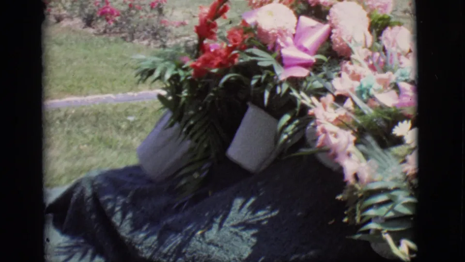 1966 Des Moines home-movie frame focused on colorful funeral flowers and greenery near the lavender casket in a grassy outdoor setting.