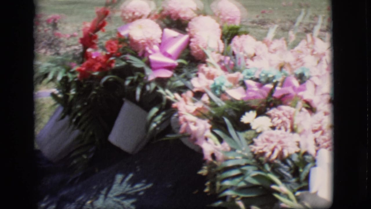 Pink and violet flowers with ribbon resting near a dark grave covering and grass.