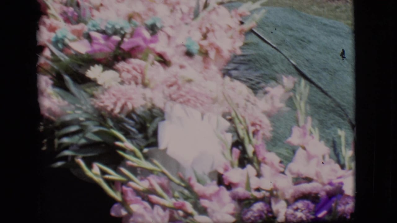 Close 8mm view of a graveside bouquet in Des Moines, Iowa, with pale pink flowers, greenery, and a dark green covering.