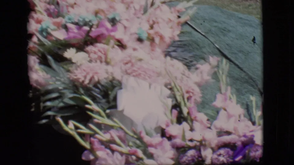 Close 8mm view of a graveside bouquet in Des Moines, Iowa, with pale pink flowers, greenery, and a dark green covering.