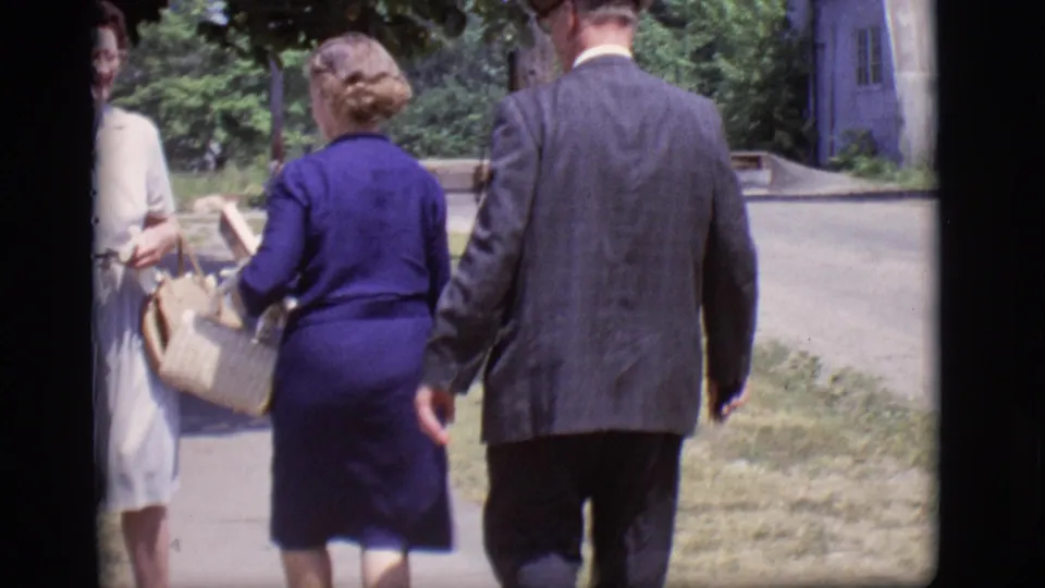 Adults in mid-century attire walk on a sidewalk in Des Moines, Iowa, with lawn and trees behind them.