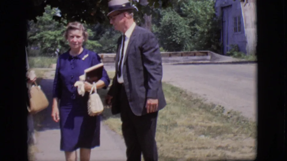 A woman holding items and a man in a suit and fedora face the camera on landscaped grounds in 1966 Des Moines footage.
