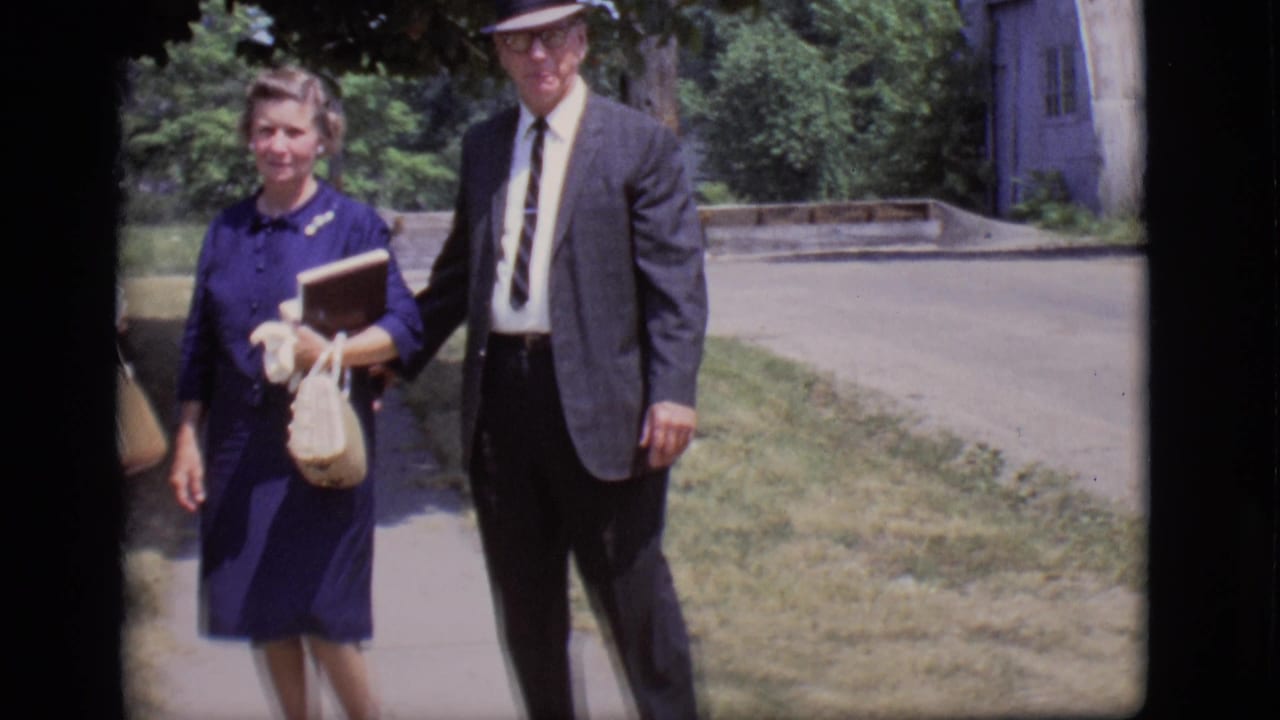 Woman in blue dress and suited man stand or walk on a sunny sidewalk.