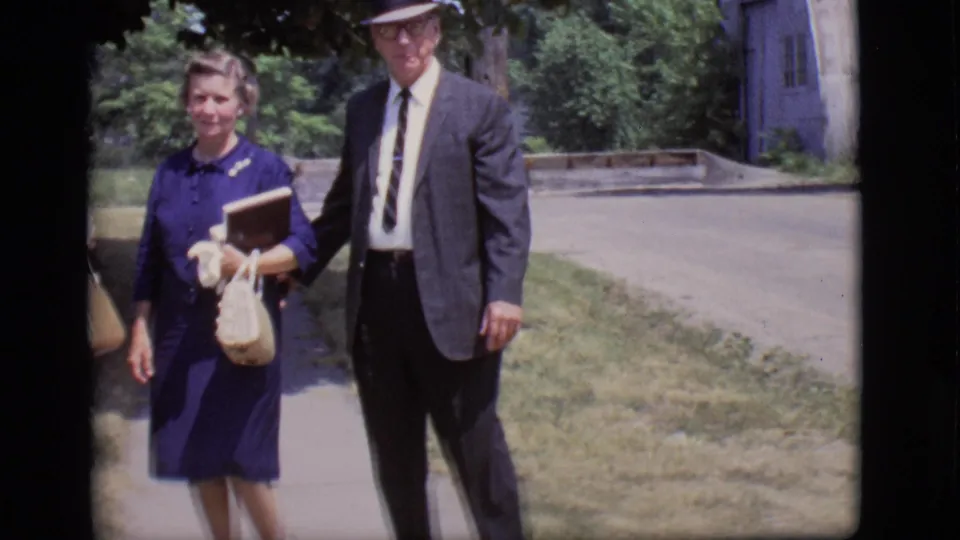 In 1966 Des Moines home-movie footage, a woman in a blue dress carries a book and purse beside a man in a dark suit and hat.