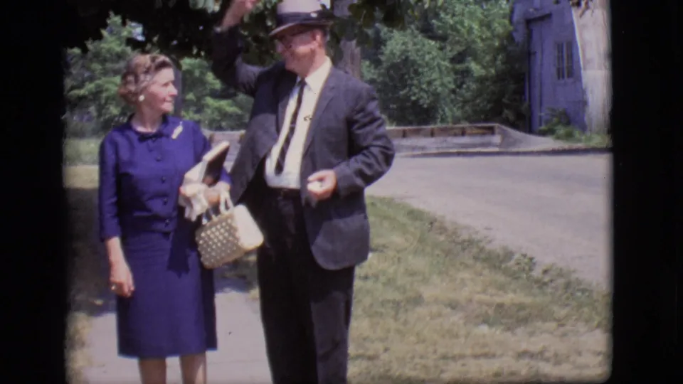 The man in a dark suit and hat makes a tipping gesture beside the woman in blue on a Des Moines sidewalk.
