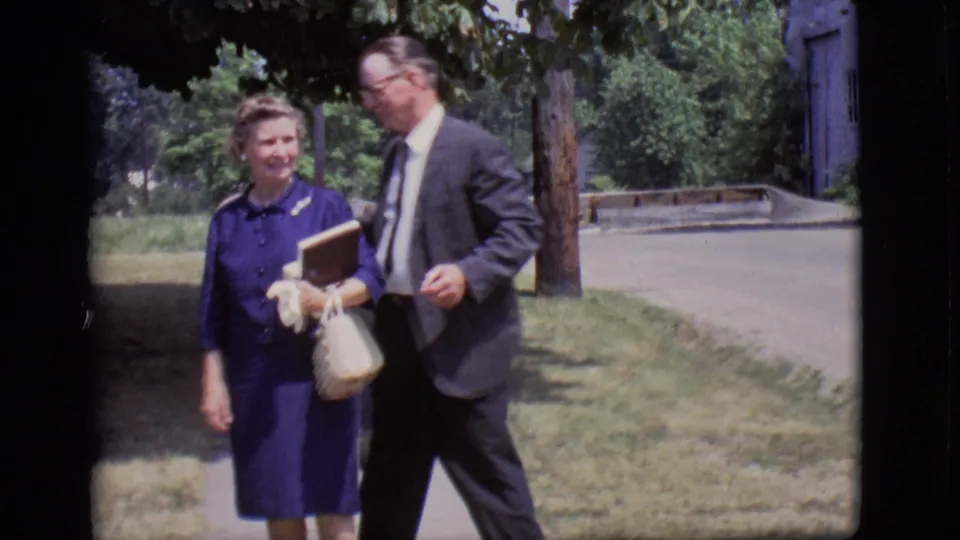 A woman carrying a book and purse walks with a suited man under tree shade in the 1966 Des Moines home movie.