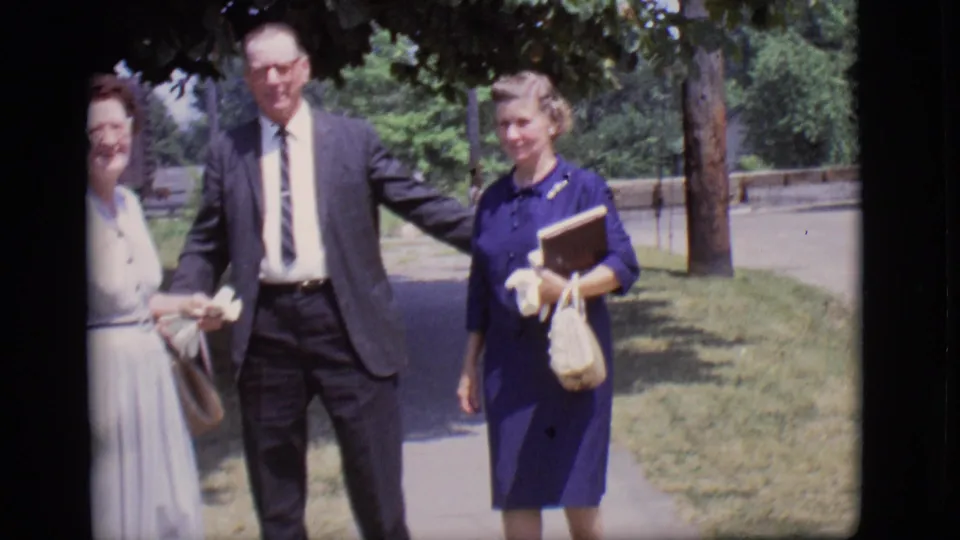 The suited man stands with a woman in white partly visible and the woman in blue holding a book in 1966 Des Moines footage.