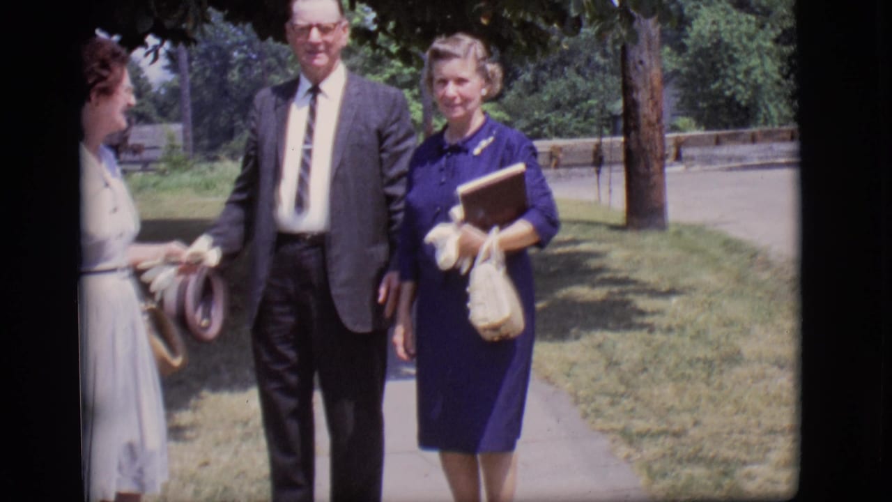Three well-dressed adults stand or walk on a sunny sidewalk under trees.